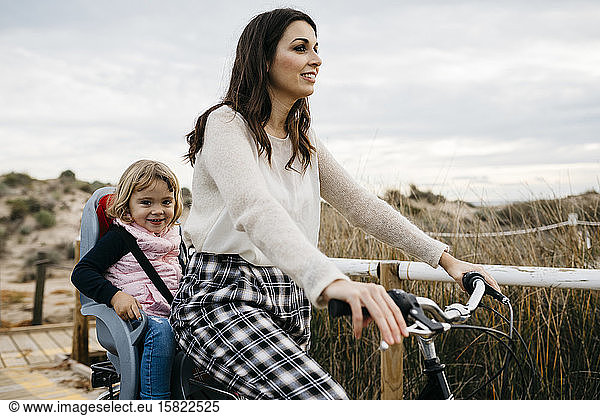 Radfahrende Frau auf einer Strandpromenade auf dem Land mit Tochter im Kindersitz
