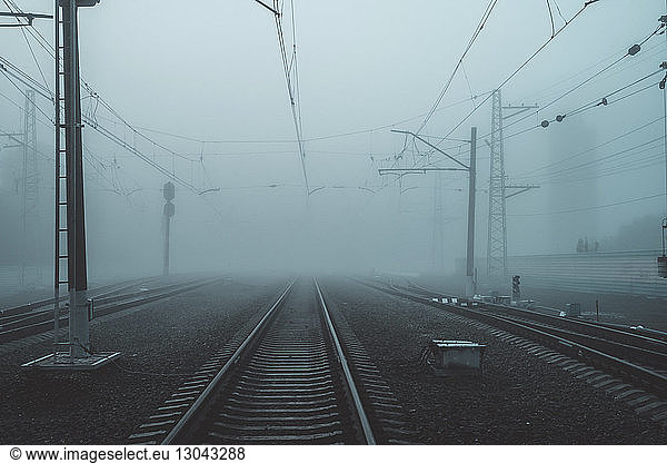 Railroad tracks by electricity pylons in city during foggy weather