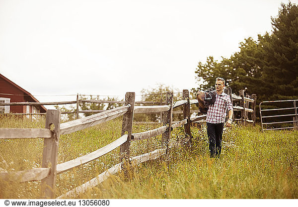 Rancher carrying saddle while walking by fence on grassy field