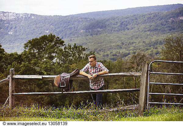 Rancher with saddle leaning on railing while standing on grassy field against mountain