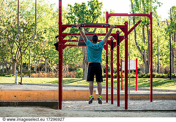 Rear view of a dark-haired athlete with beard hanging from a calisthenics bar in a park. . Outdoor crossfit concept.