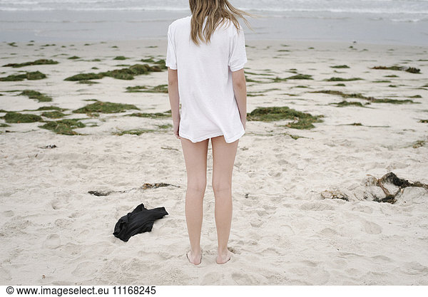 Rear view of a girl with blond hair in a white t-shirt  standing on a sandy beach.