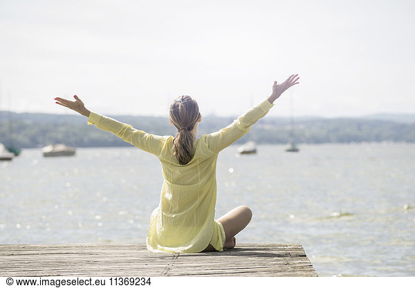 Rear view of a woman with arms raised sitting on boardwalk at the lake  Ammersee  Upper Bavaria  Germany
