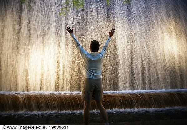 Rear view of a woman with her hands raised in front of a waterfall fountain.