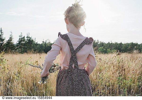rear view of a young girl walking through a meadow holding wildflowers