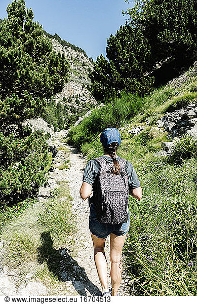 Rear view of anonymous female traveler in casual clothes with a backpack walking along a mountain path in a sunny day