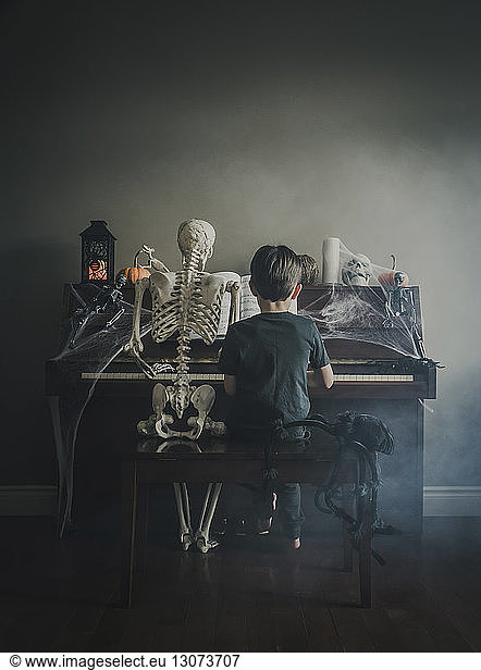Rear view of boy playing piano while sitting by skeleton during Halloween