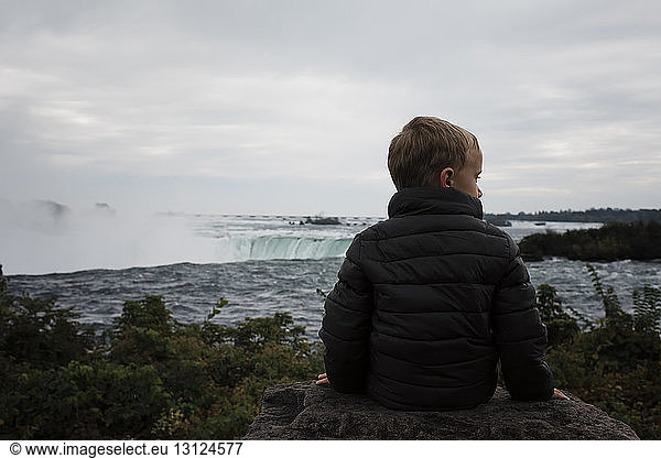 Rear view of boy sitting on rock while sitting against Niagara Falls