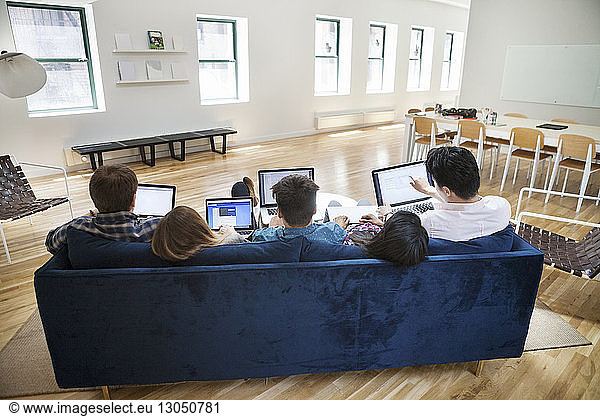 Rear view of business people using laptops on sofa in creative office