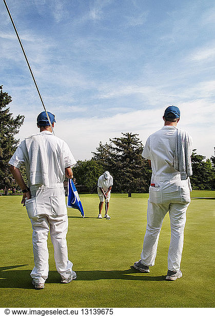 Rear view of caddies looking at golfer taking a shot in golf course