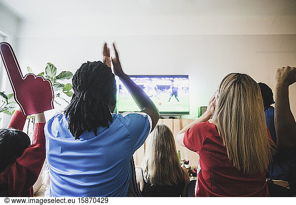 Rear view of excited soccer fans watching match with friends at home