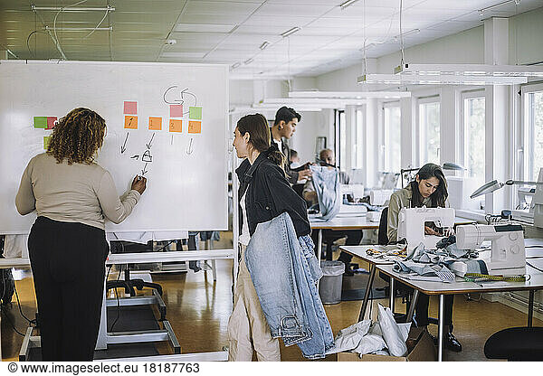 Rear view of fashion designer explaining female colleague while writing on whiteboard at workshop