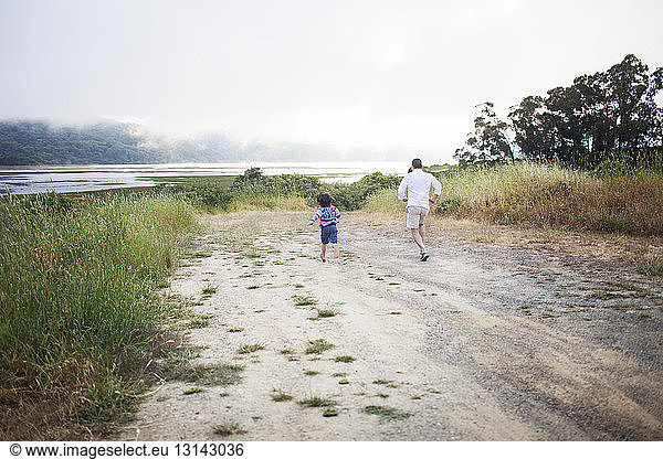 Rear view of father and son running on dirt road against sky