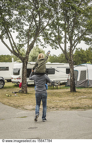 Rear view of father carrying girl on shoulders while standing against camper van