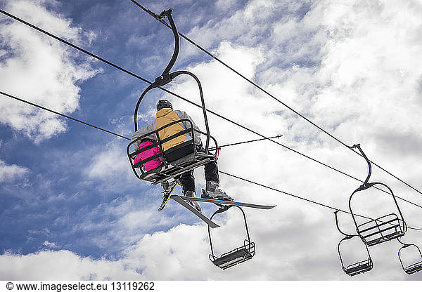 Rear view of father with daughter sitting on ski lift against cloudy sky during winter