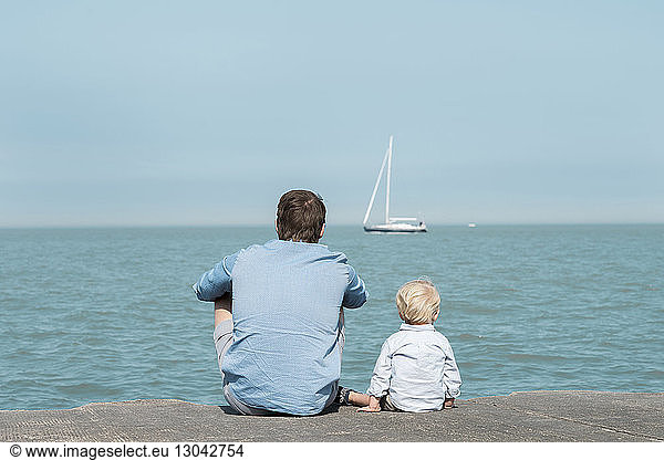 Rear view of father with son sitting at beach against blue sky during sunny day, Rear view of father with son sitting at beach against blue sky during sunny day