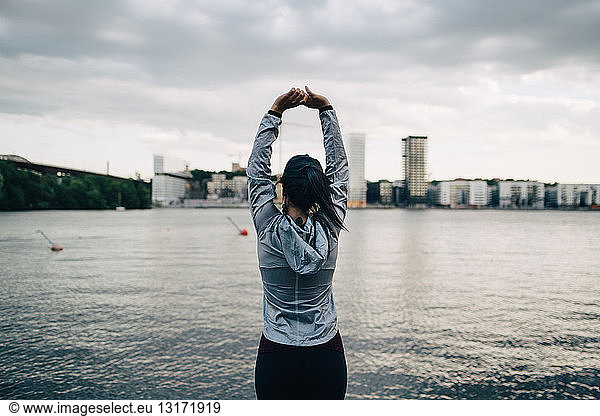 Rear view of female athlete stretching arms while standing against sea in city