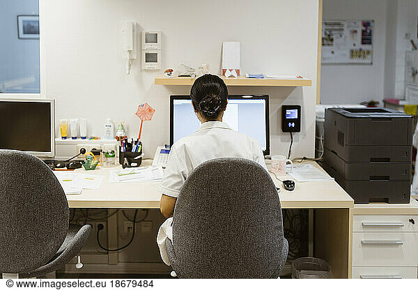 Rear view of female doctor working on desktop computer while sitting on chair at clinic