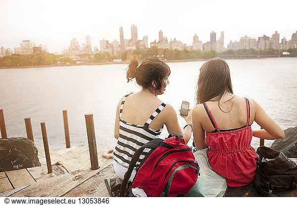Rear view of female friends using phone while sitting at riverbank