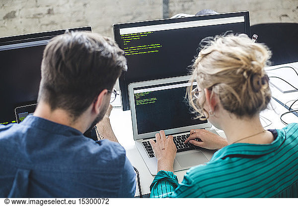 Rear view of female IT expert coding in laptop while sitting by male hacker at workplace