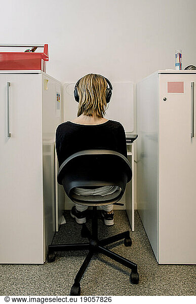 Rear view of female teenage student wearing headphones sitting on chair in library at high school