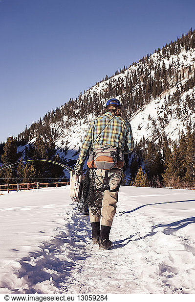 Rear view of fly fisherman walking on snowy field against mountain