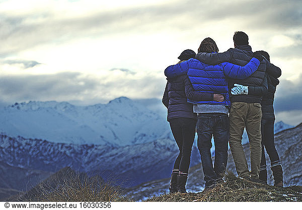 Rear view of four people standing arm in arm on a mountain  snow-capped peaks in the distance.