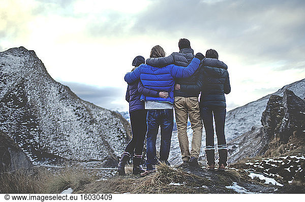Rear view of four people standing arm in arm on a mountain  snow-capped peaks in the distance.