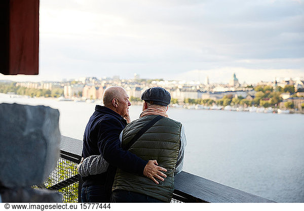 Rear view of gay couple with arm around standing against railing by river in city