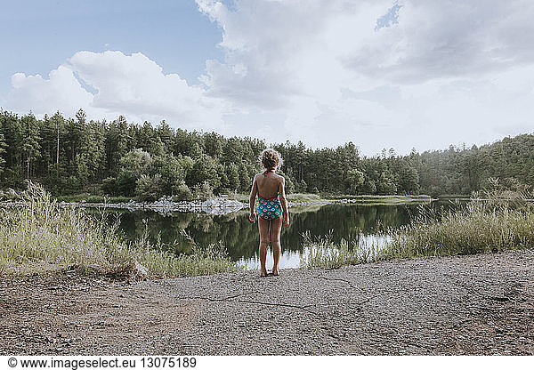 Rear view of girl in swimwear standing at lakeshore against cloudy sky