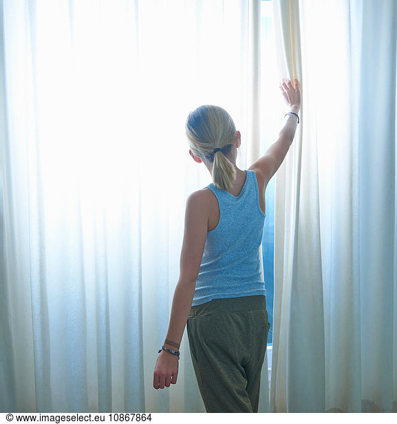 Rear view of girl peering through net curtained window