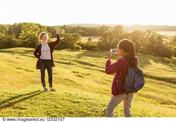Rear view of girl photographing happy mother showing peace sign on field against clear sky during sunset