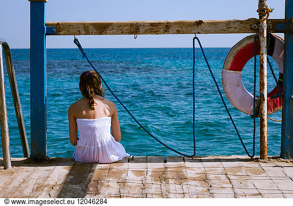 Rear view of girl sitting on pier looking out to sea