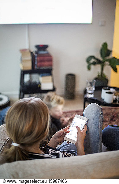 Rear view of girl using smart phone while sitting on sofa at home