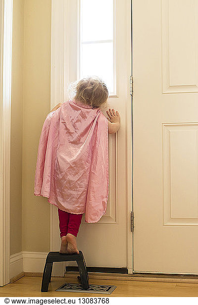 Rear view of girl wearing cape standing on stool by door at home