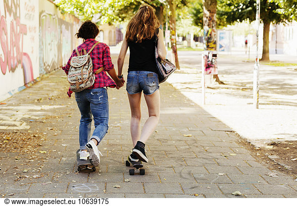 Rear view of girls holding hands and skateboarding on footpath