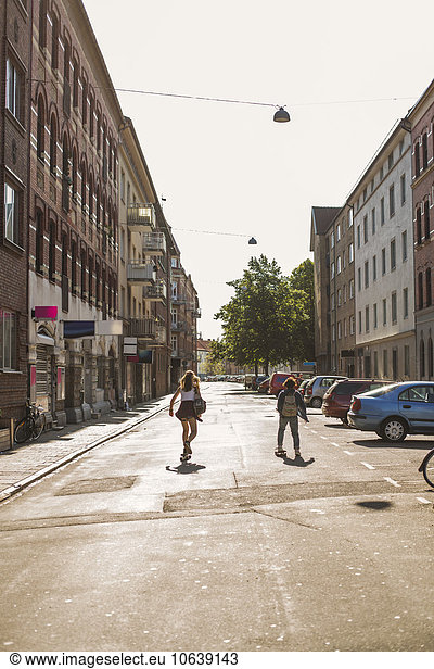 Rear view of girls skateboarding on street amidst buildings