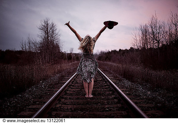Rear view of happy woman with arms raised standing at railroad tracks against cloudy sky