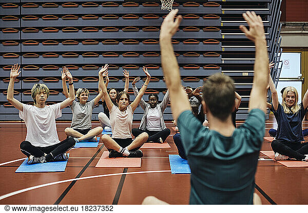 Rear view of male coach with arms raised teaching yoga to students in sports court