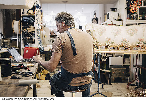 Rear view of male upholstery worker using laptop at workbench in workshop
