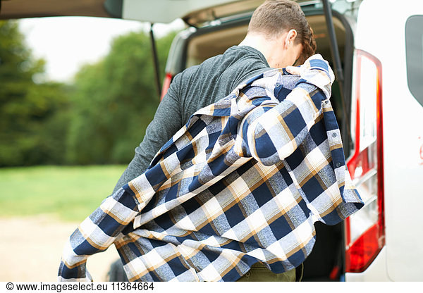 Rear view of man by automobile boot putting on plaid shirt