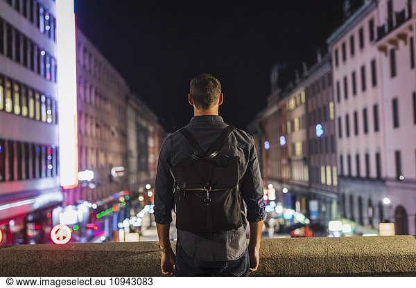 Rear view of man carrying backpack standing on bridge in city at night