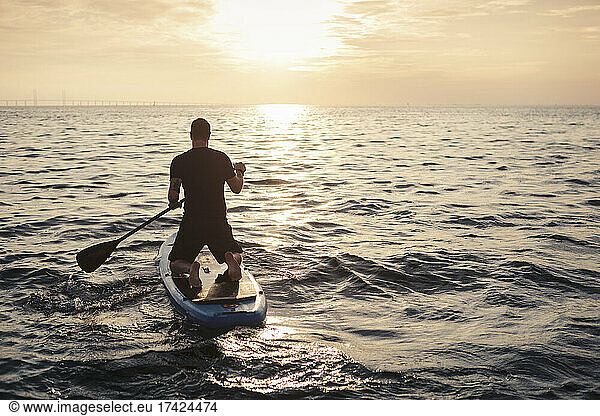 Rear View of man rowing paddleboard in sea during sunset