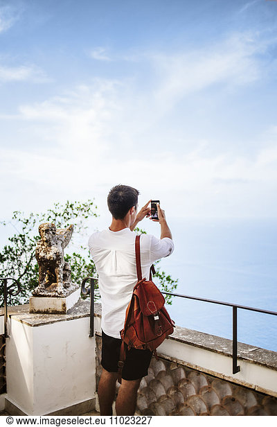 Rear view of man standing at railing and clicking selfie against sky