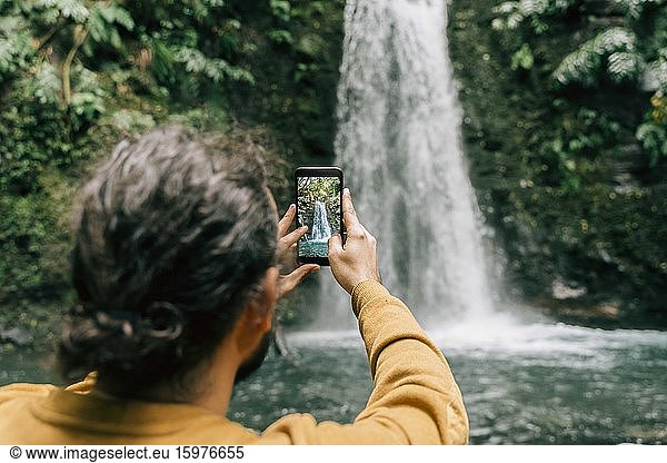 Rear view of man taking smartphone picture at a waterfall on Sao Miguel Island  Azores  Portugal