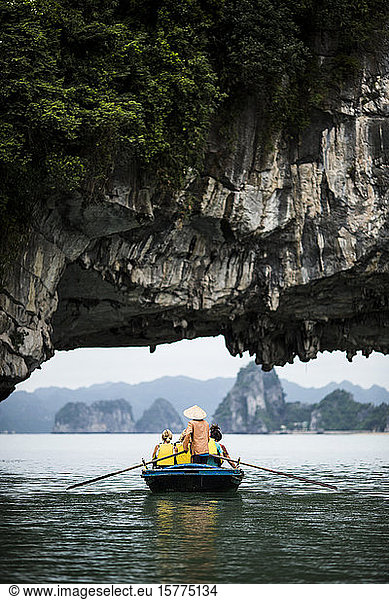 Rear view of man wearing straw hat transporting small group of people on a boat  rowing underneath natural rock arch.