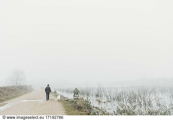 Rear view of man with dog walking by lake in fog