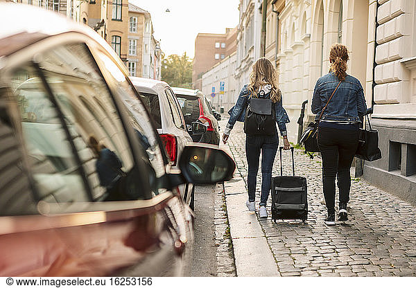 Rear view of mother and daughter with suitcase walking at roadside during weekend