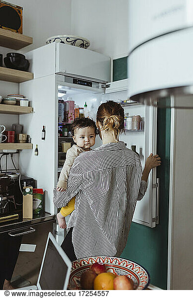 Rear view of mother carrying baby boy while opening refrigerator in kitchen