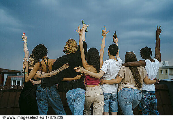 Rear view of multiracial young men and women gesturing with arms raised on rooftop at dusk